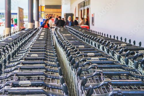 Rows of shopping cart lined at supermarket entrance with customers in background near retail store. Shopping cart usage in grocery retail, consumerism purchase, store service