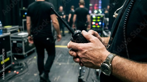 Close-up on a security guard's hand holding a walkie-talkie in a backstage area with other people and equipment in the background, suggesting event security or crowd control operations