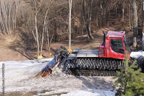Snowcat working on maintaining the ice-covered trail in a forest area. The image shows the red snowcat machine operating on a partially snowy path, surrounded by bare trees.