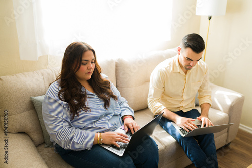 Business partners concentrating on their laptops, sitting apart on a sofa while working remotely from their cozy home