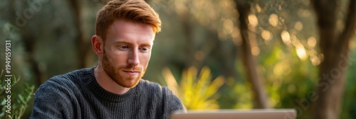 Caucasian male young adult redhead laptop outdoors in warm garden light by olive trees wearing knit sweater focused on work and reading screen