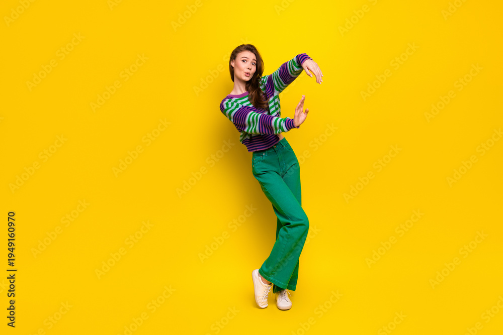 Obraz premium Young female model dances in a colorful striped sweater and green pants against a bright yellow background