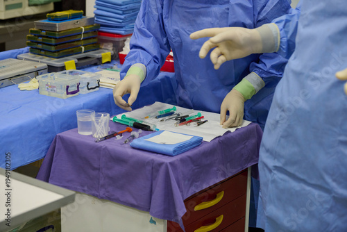 Fotografie Surgeons in blue sterile gowns and latex gloves with green cuffs preparing instruments: syringes, scalpels, forceps on purple drape in operating room