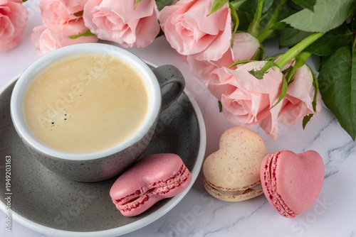 Vintage granite color coffee cup, pink rose flowers and french sweet cookies macarons macaroons on marble background. Heart shape macarons, cup and bouquet of roses.