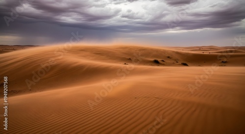 A dramatic desert scene of sand dunes under a stormy, overcast sky. Sand is blowing