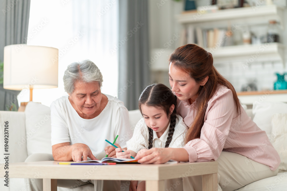 Fototapeta premium Asian grandmother and mother lean in together to help their young granddaughter with a coloring book at a living room table — a gentle three-generation learning moment.