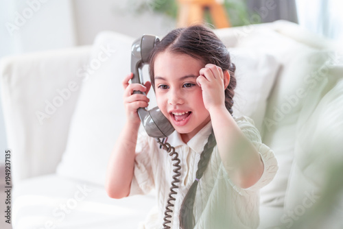 Εκτύπωση καμβά Delightful young Asian girl bursts into laughter while holding a vintage corded telephone receiver on a white sofa, capturing pure childhood joy and spontaneity