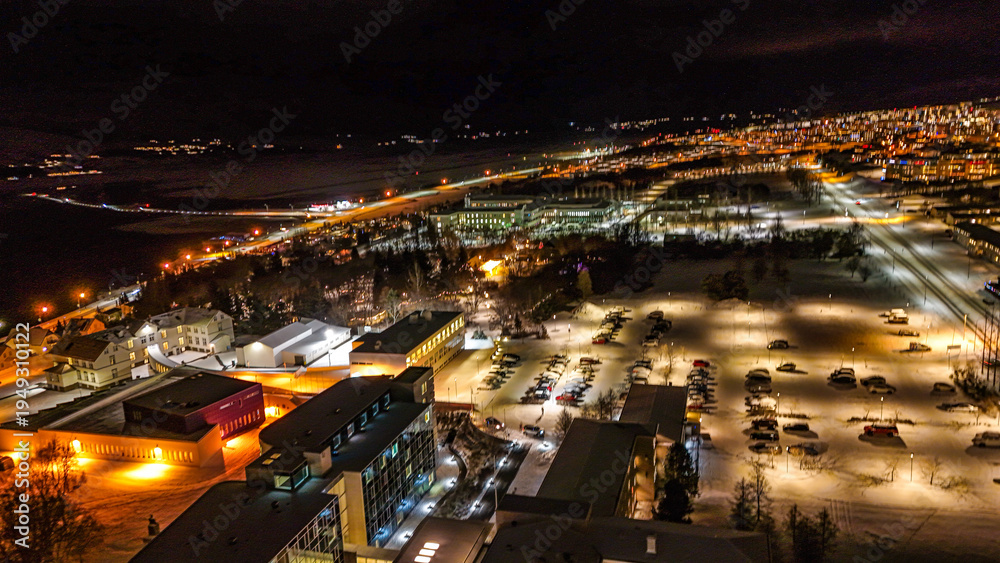 Fototapeta premium Drone view of Akureyri in North Iceland at night with illuminated bridge and city lights reflecting on water. Beautiful aerial scene of Akureyri harbor showing urban landscape and coastline in Iceland