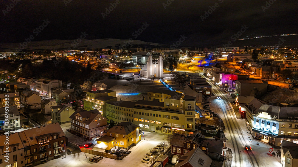 Fototapeta premium Drone view of Akureyri in North Iceland at night with illuminated bridge and city lights reflecting on water. Beautiful aerial scene of Akureyri harbor showing urban landscape and coastline in Iceland
