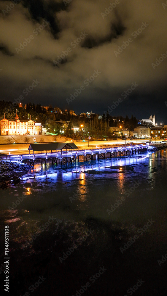 Fototapeta premium Drone view of Akureyri in North Iceland at night with illuminated bridge and city lights reflecting on water. Beautiful aerial scene of Akureyri harbor showing urban landscape and coastline in Iceland