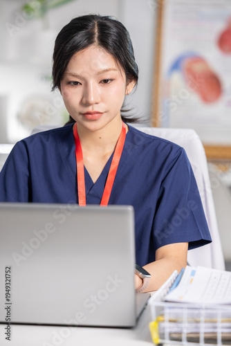 Young Asian female nurse wearing navy blue scrub looking heavily focused and serious while typing on silver laptop computer sitting at modern medical clinic desk reviewing patient health record
