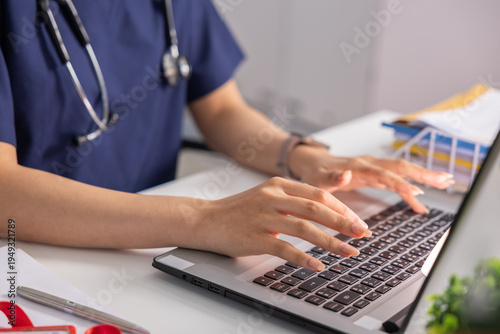 Medical professional in blue scrub with stethoscope typing carefully on laptop keyboard at clinic desk showing calm focused dedication to healthcare patient record management