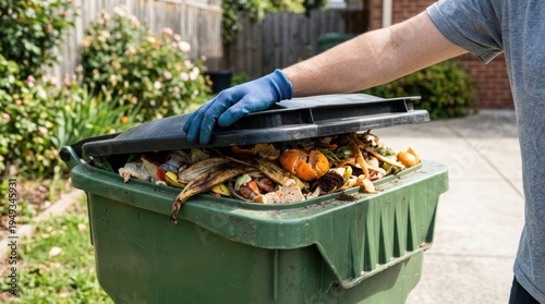 A person wearing gloves is disposing of food waste into a green recycling bin.