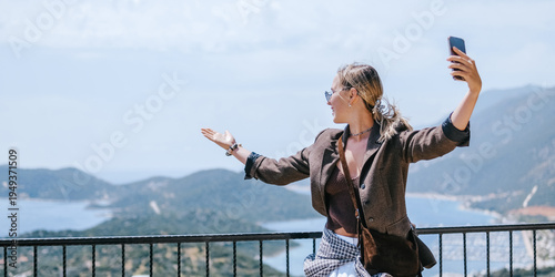 Travel blogger filming live stream on smartphone against mountain coastline, Gen Z female tourist on viewpoint, panoramic sea and hills, social media content creator, sunny outdoor vacation vibe.