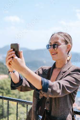 Travel blogger filming live stream on smartphone against mountain coastline, Gen Z female tourist on viewpoint, panoramic sea and hills, social media content creator, sunny outdoor vacation vibe.