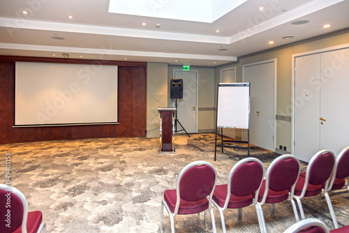 Empty corporate conference room with large projection screen, flipchart and rows of red chairs set up for a business presentation, seminar or training event in a modern meeting space.