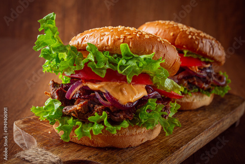 Tablou pe pânză Two burgers with torn beef, tomatoes, fried onions and sauce, laid out on a wooden board, there are no people