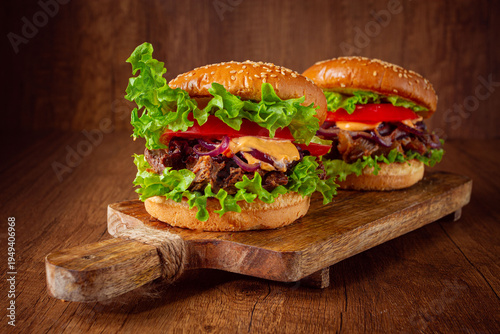 Billede på lærred Two burgers with torn beef, tomatoes, fried onions and sauce, laid out on a wooden board, there are no people
