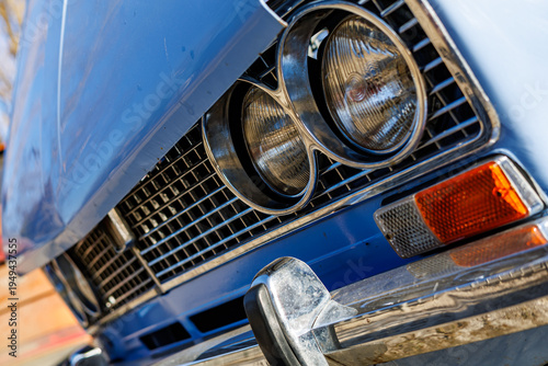 Fotografie Close-up of a blue vintage car front: headlights, chrome grille and retro whitewall tires on a cobblestone road