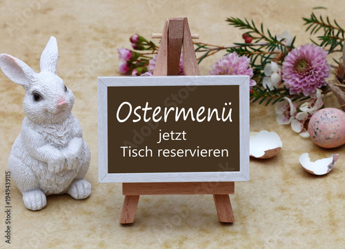 Holzstaffelei mit dem Text Ostermenü jetzt Tisch reservieren, neben weißem Osterhasen und Frühlingsblumen mit Eierschalen auf hellem Untergrund.