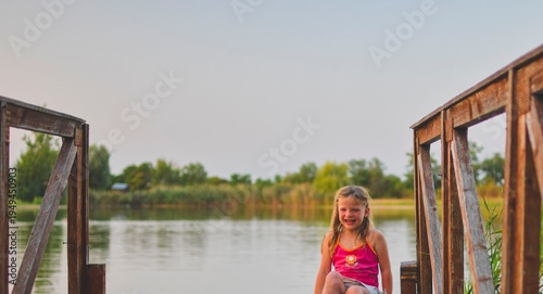 A lovely little girl sits on a wooden pier at the shore of a lake. The photo was taken around sunset, during the golden hour. It evokes childhood, summer holidays, looking at