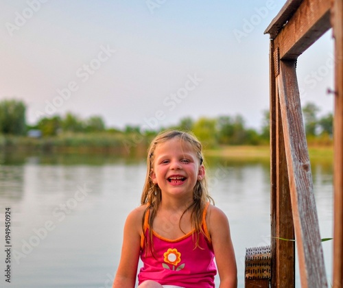 A cute little girl with a smile on her face sits on a wooden pier at the side of a lake. The photo was taken during the golden hour, at sundown. It evokes the concept of childhood, summer 