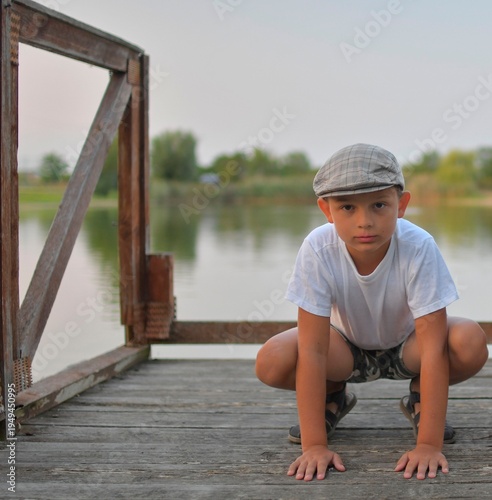 A cute little boy wearing a flat cap sits on a wooden pier by the lake. Small kid sits on a jetty by the edge of a pond. The concept of carefree childhood and summer holidays.