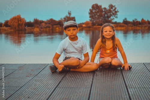 A group of cute little children sit on a wooden pier by a lake. Small children looking into the future. Children sit on a dock. Siblings. Two children of different ages