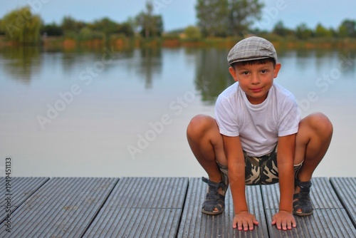 A cute little boy sits on a wood pier by a lake. A young boy wearing a flat cap looks into the future. A small child sitting on a pier. A school-aged boy sits on a wooden pier by a pond. 