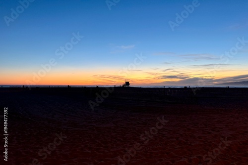 Silhouette of a lifeguard tower at Santa Monica Beach after sunset, glowing evening sky and warm holiday atmosphere over the calm Pacific coast.