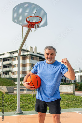 vertical Senior man smiling at camera holding a basketball and flexing his bicep on an outdoor court, symbolizing vitality, wellbeing, and active aging in retirement