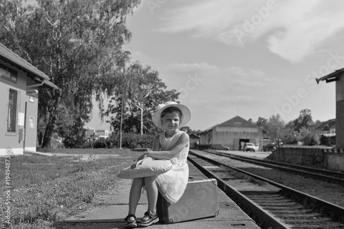 Young girl in a hat and dress sits on a suitcase at a train station platform with tracks