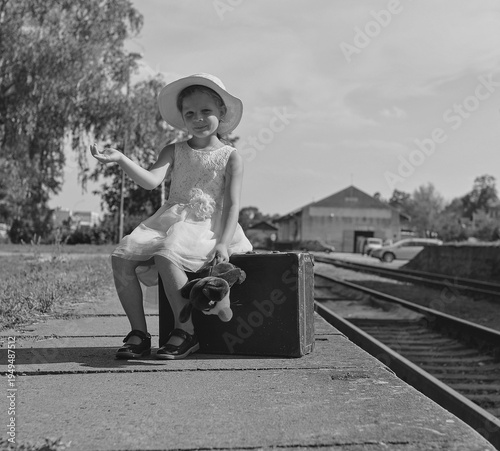 Young girl in a hat and dress with a suitcase and toy at a train station platform