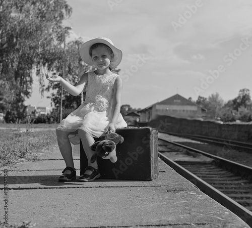 Young girl in a hat and dress sitting on a suitcase at a train station with her toy