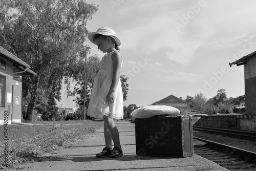 Young girl in a dress and hat waits with a suitcase on a train platform