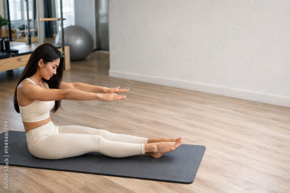 Obraz premium Young woman exercising on yoga mat in fitness studio stretching her arms forward with legs extended, focused on wellness and flexibility training