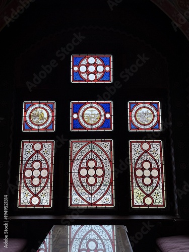 Illuminated stained glass window with red, blue, and white geometric motifs glowing in a dark historic interior, creating a dramatic medieval church atmosphere.
