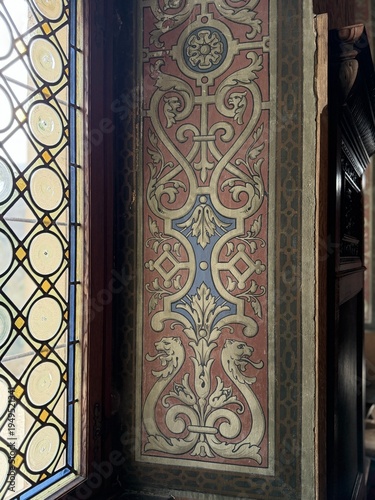 Ornate historic wall panel beside stained glass window, featuring elegant scrollwork, floral motifs, and heraldic animal details in a richly decorated heritage interior.