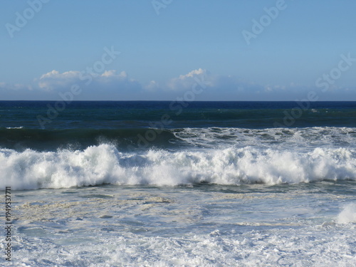 Powerful ocean waves crash along the coastline in Morocco, creating white sea foam as they roll toward the shore. The deep blue Atlantic Ocean stretches toward the horizon under a clear sky with a few
