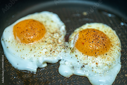 Two sunny side up fried eggs in a pan seasoned with black pepper and spices. Close-up of fresh organic breakfast cooking. Healthy protein meal background