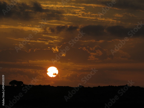 A dramatic sunset landscape captured in Morocco, where the glowing orange sun sits low on the horizon beneath layers of dark clouds. The sky is filled with warm golden and amber tones, creating a stri