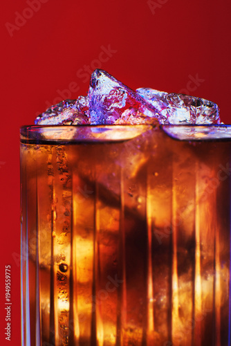 Macro photo of a glass with a carbonated drink and ice cubes. Contrasting blue and purple neon lights on the ice on a rich red background. 