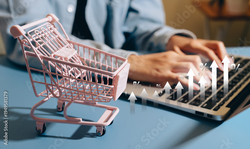 Ecommerce growth concept, miniature shopping cart on desk with laptop and rising percentage arrows, online retail expansion, digital sales increase, consumer demand trend