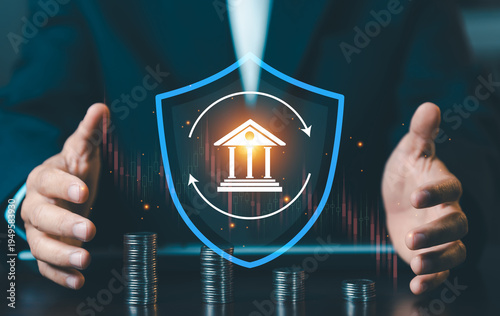 Businessman protecting bank icon inside blue shield with stacks of coins, financial insurance and capital protection concept against market volatility.