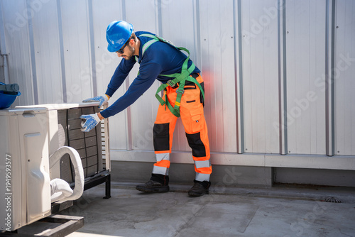 Technician in safety harness installing an outdoor air conditioning unit, Maintenance worker repairing HVAC system on a roof, Male engineer inspecting industrial air conditioner condenser outdoors