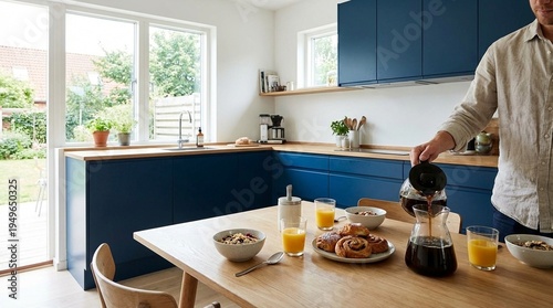 Bright modern kitchen with blue cabinets and wooden table, person pouring coffee beside breakfast pastries and orange juice, calm morning routine at home