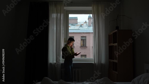 Upset shocked senior woman reading news or checking message on smartphone online, lady standing in dark bedroom in front of window with phone, elderly female with sad expression scrolls phone sighing.