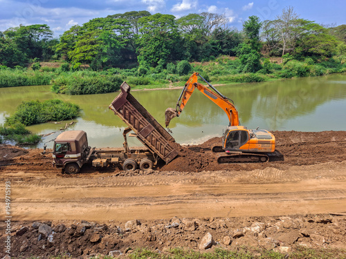 Construction work is taking place by the river; trucks are lifting their trailers to unload soil, and an orange backhoe is parked nearby, assisting in clearing the earth.