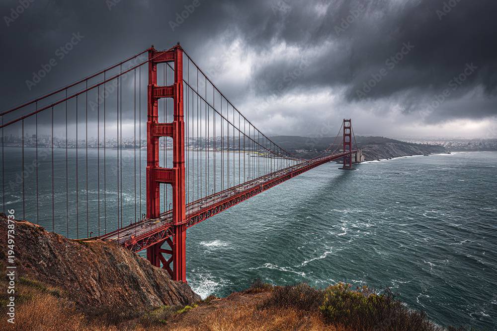 Fototapeta premium Golden Gate Bridge suspension structure over turbulent water under dramatic stormy sky with rocky foreground and distant cityscape on horizon
