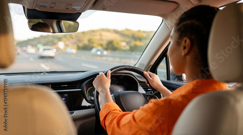 Photos Rear view of a focused African American woman confidently driving a modern car on a busy highway during a bright